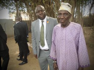 A man standing with the elderly caretaker of Opa Oranmiyan, a direct descendant of Oranmiyan, beneath large trees at the site.