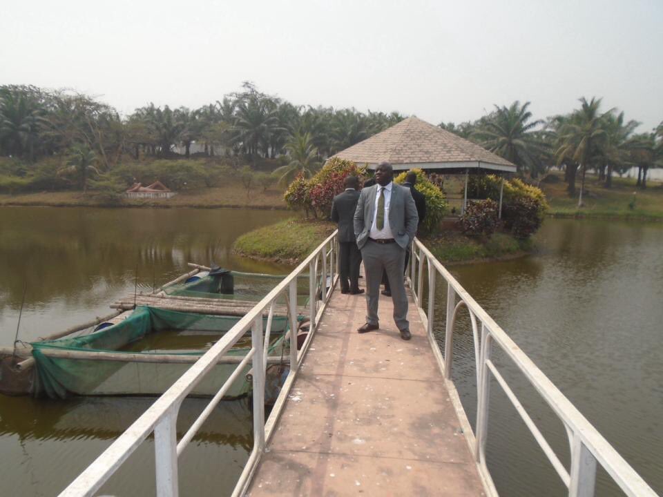 A man in a grey suit standing on a bridge over the private lake at Chief Duduyemi's house in Ile Ife, with a gazebo and fish cages visible on the water.