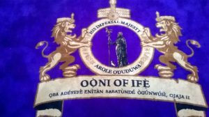 The royal crest of the Ooni of Ife, featuring two lions holding a shield with a crown, displayed on purple fabric.