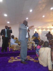Bayo Adeyinka speaking to a seated crowd of farmers inside the palace hall, with the Ooni's crest visible on the carpet.