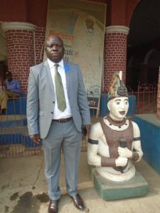 Bayo Adeyinka standing beside a statue of an Emese, a palace attendant of the Ooni, near a banner marking a royal birthday celebration.